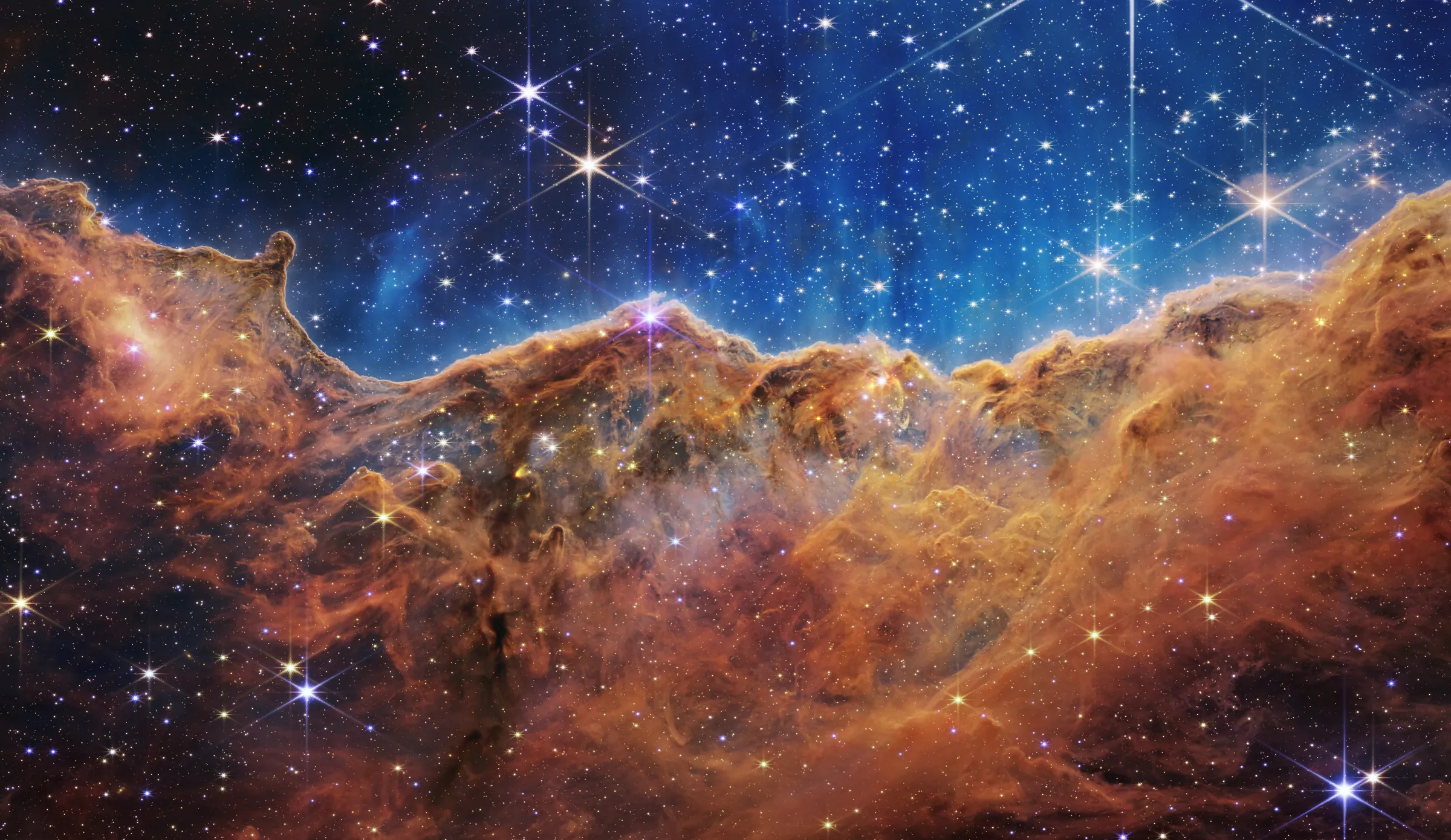 The edge of the star-forming region NGC 3324 in the Carina Nebula, a wall of gas and dust under a field of stars, seen in infrared light by the James Webb Space Telescope.