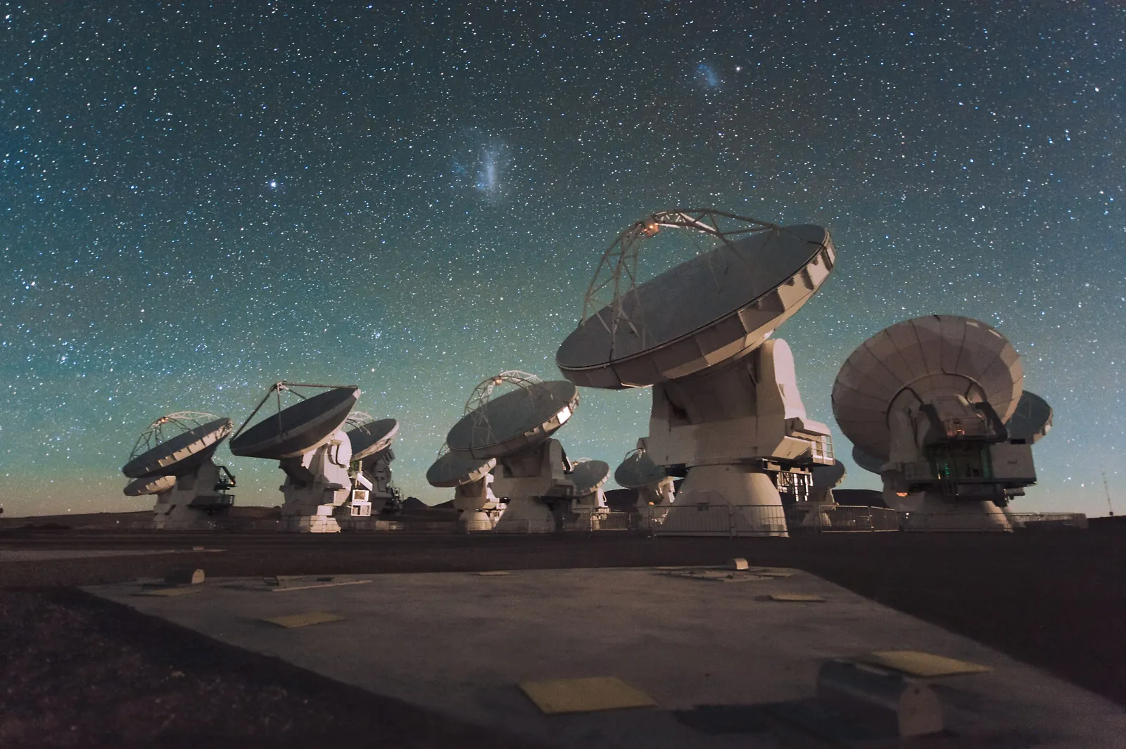 A group of ALMA radio antennas observing at night on the Chajnantor plateau, with the Large and Small Magellanic Clouds in the sky above.